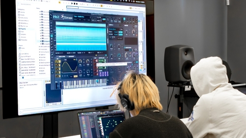 Two students sit at desks with their laptops. In front of them, a large screen shows a digital music mixing platform.