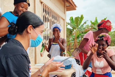 A student wearing a surgical mask smiles and claps with a group of Ghanaian women