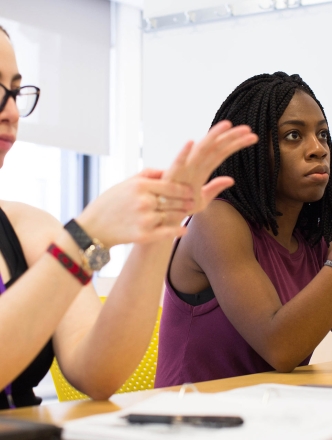 Two students holding one of their hands while seated at a table in a classroom.